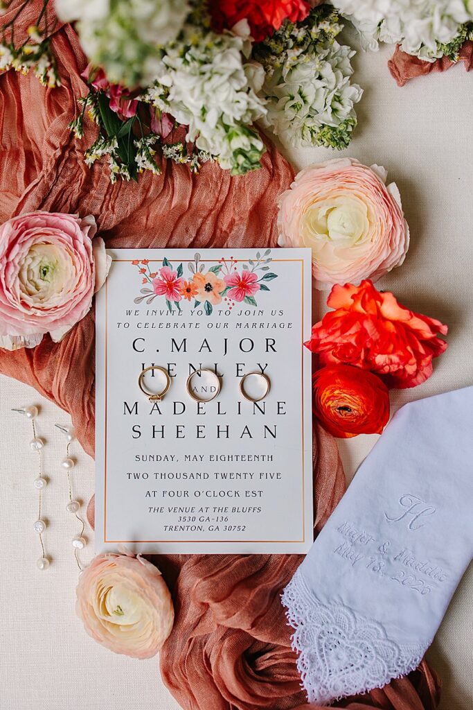 flat lay shot of wedding invitation, wedding rings, pearl earrings, handkerchief and ranunculus