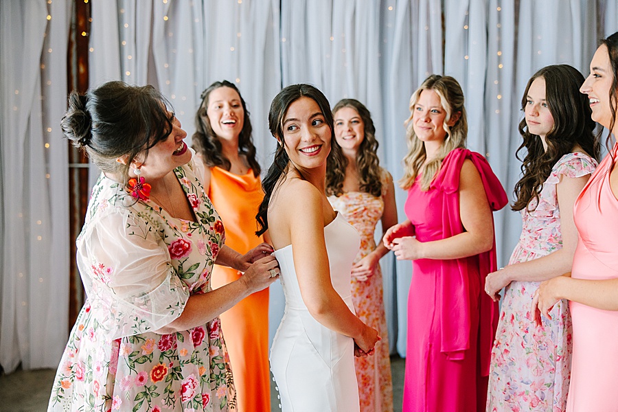 bridesmaids looking on bride while mom helps her to get dressed on her wedding day