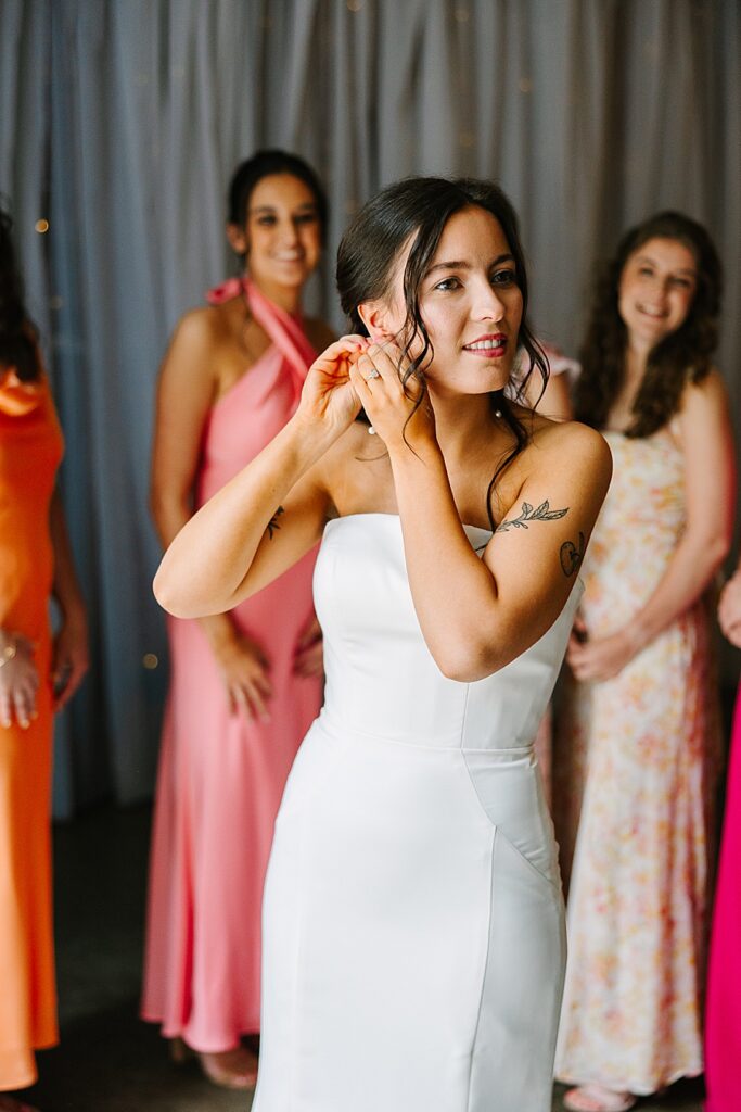 bride putting on earrings on wedding day with bridesmaids watching in the background