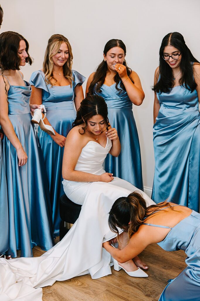 Bridemaids in blue satin dresses helping Bride put on her shoes in bridal house at the Glass house at Nichols Heir