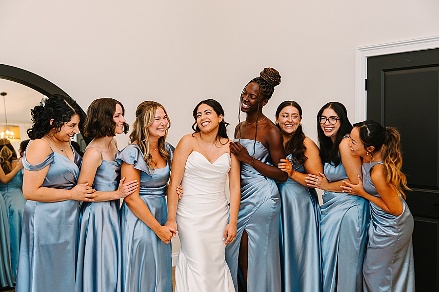 Bride laughing with bridesmaids in blue satin dresses at the Glass House at Nichols Heir bridal house while getting ready for wedding day
