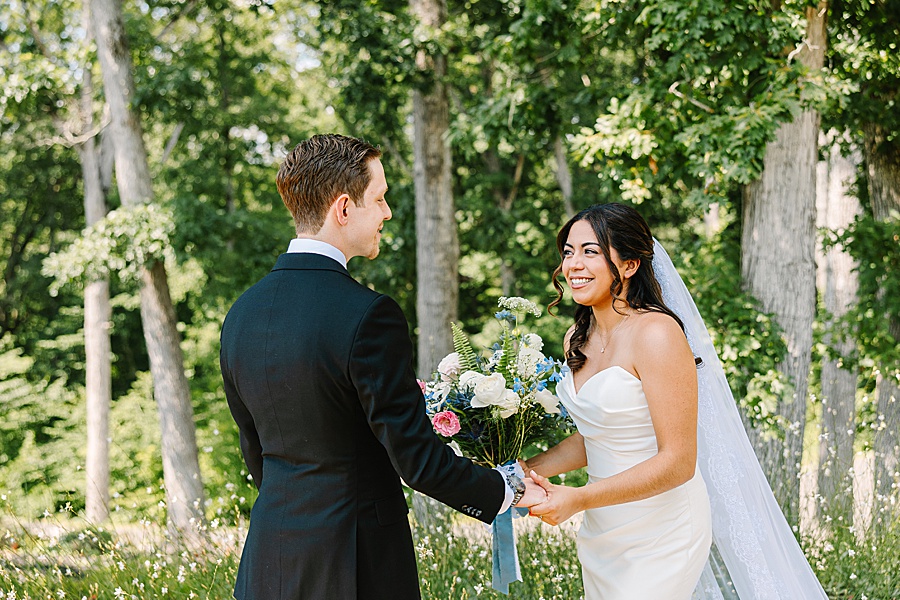 Bride and groom holding hands during first look on wedding day with bouquet of lush florals of delphinium, Ranunculus, and garden roses