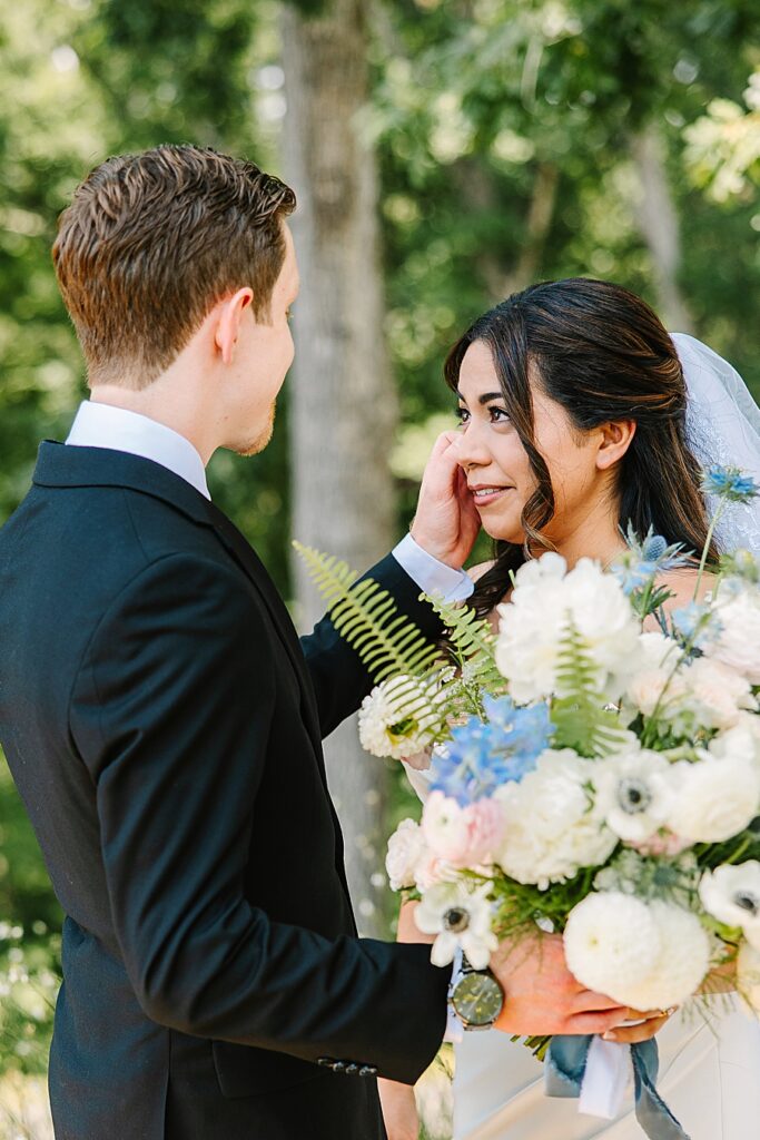 groom brushing away tear from bride on wedding day with lush floral bouquet in image with ferns, delphinium, white poppies, pink Ranunculus